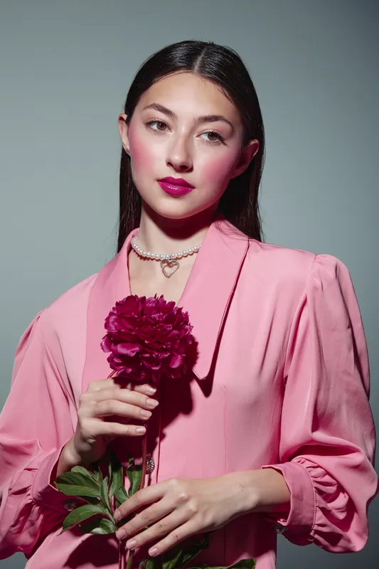 Young woman in pink elegant blouse holds dark red flower while posing with stylish makeup and flawless skin on neutral background. Concept for beauty, creative fashion, and self-expression.