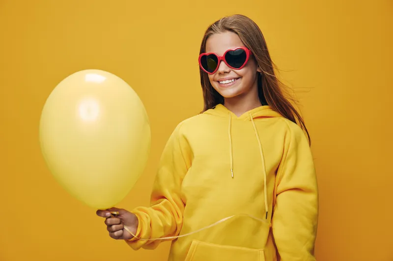 Girl in yellow hoodie and heart-shaped sunglasses holding yellow balloon smiling on bright yellow background with casual style and happy expression in studio lighting.