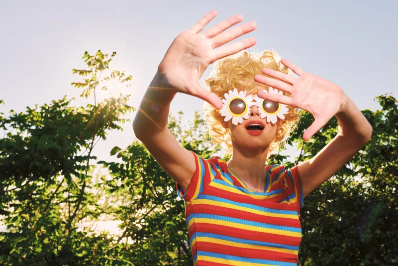 Playful woman wearing a striped top and whimsical flower glasses outdoors, sunlit garden backdrop, bright colors and retro vibe evoke carefree self expression and wellness in a candid lifestyle scene.