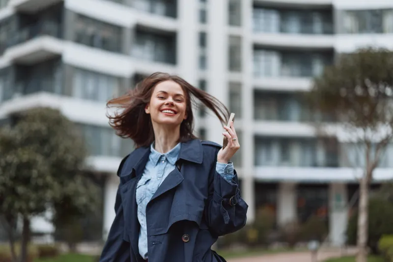 Young woman with shoulder-length brown hair smiles joyfully while walking outdoors in a blue trench coat and light blue shirt in an urban environment.