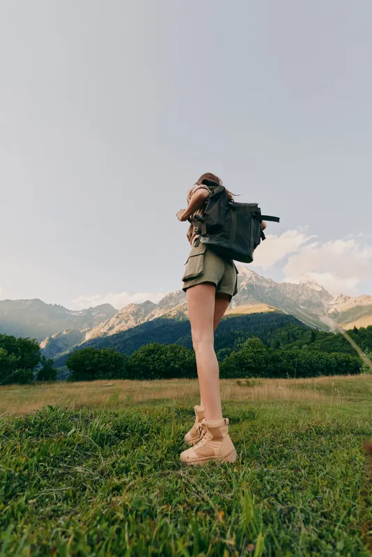 Young woman with backpack hiking in mountains during daytime. She stands on green grass with clear sky and mountain peaks in the background. Outdoor lifestyle concept.
