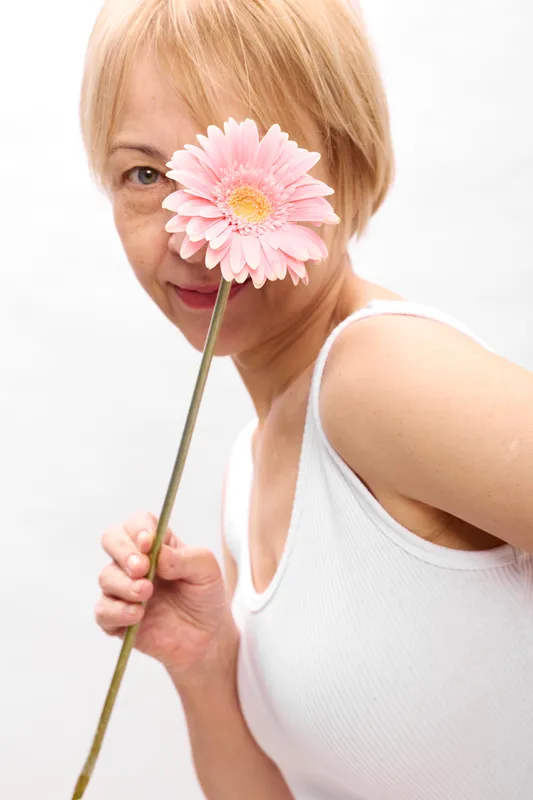 Blonde woman holding pink flower by her face on white background, soft studio portrait lighting, smile, calm beauty and spring freshness, feminine gentle expression