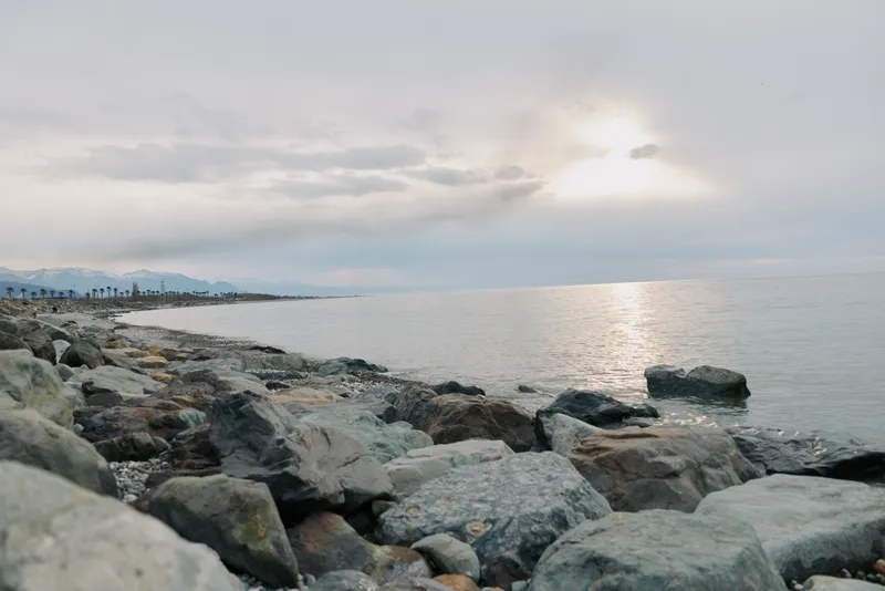 Rocky coastline with calm sea reflecting soft light under cloudy sky. Mountains and distant shore visible in the background. Peaceful nature scene with muted colors and natural textures.