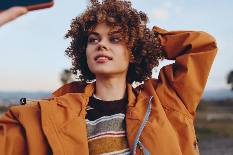 Woman lifestyle outdoors, curly hair framing her face, she wears orange jacket and knit sweater, eyes looking forward, wind and soft light, distant sky, nature background, candid portrait