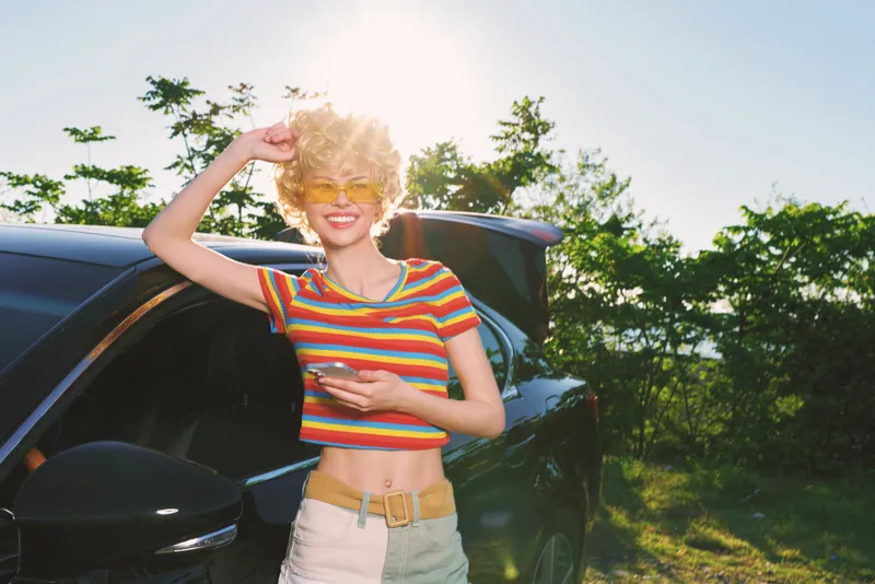 Young woman wearing a striped crop top leans against a dark car in bright sunlight, smiling with carefree energy in a vivid outdoor setting