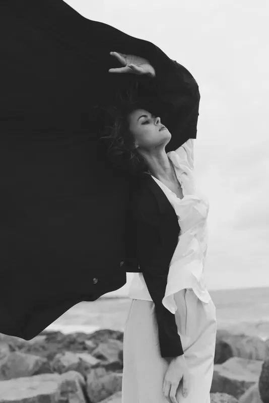 Cape, wind, and flowing dress create a dramatic beach scene with a strong silhouette, cinematic mood, and elegant contemporary fashion aesthetic editorial mood.