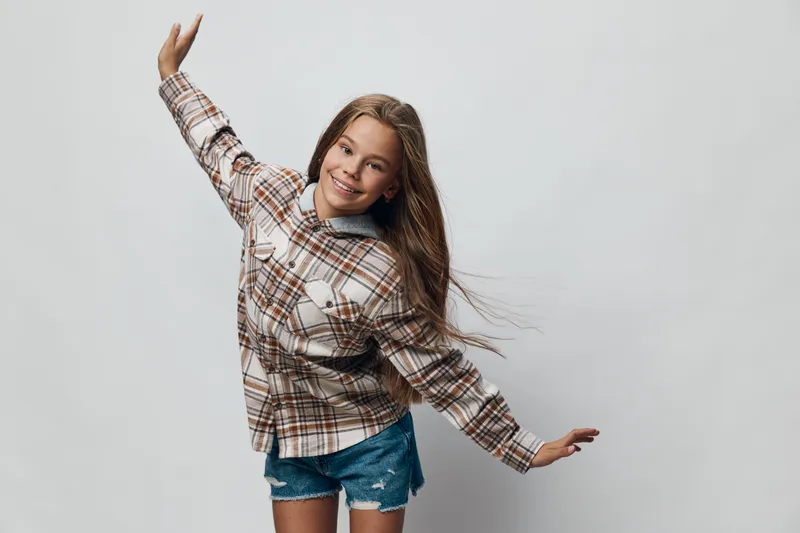Girl with long hair wearing checkered shirt and denim shorts posing with arms outstretched on plain background. Smiling child with casual outfit and natural expression for lifestyle.