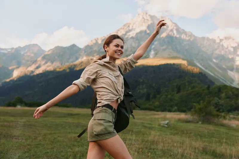 Young adult woman with a backpack stretches arms and smiles in a sunlit meadow, with snow capped mountains rising behind, conveying freedom and adventure in nature.