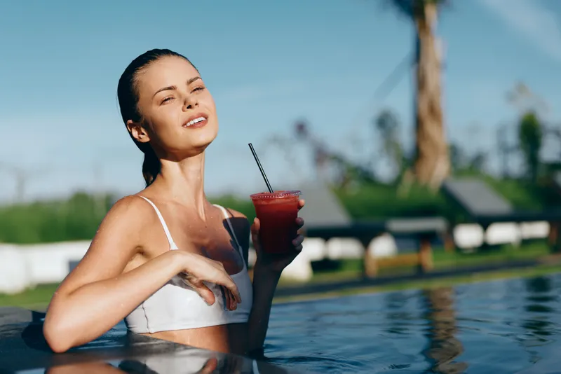 Relaxed woman in a white bikini sits in a pool, sipping a red juice through a straw, enjoying sunshine and breeze, bright outdoor scene with palm trees and blue sky.