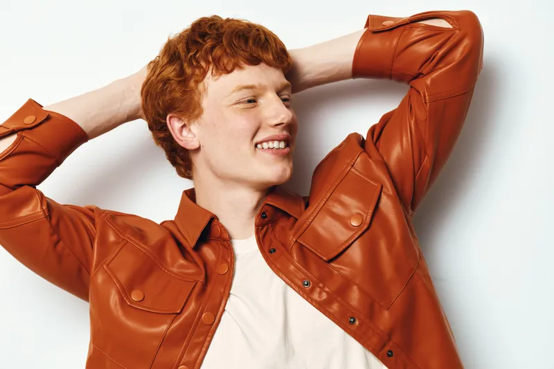 Redhead young man wearing brown leather jacket smiling and posing with hands behind head looking relaxed and confident against white background in studio.