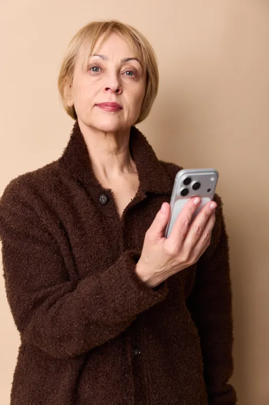 Older woman smartphone portrait indoors, casual coat on beige background, holding a mobile phone, calm expression, looking at camera, soft light, minimal composition