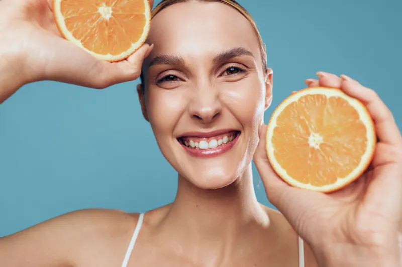 Smiling woman holding two halves of fresh orange close to face, natural skin, bright background, healthy lifestyle, cheerful expression, summer mood, vitamin C concept