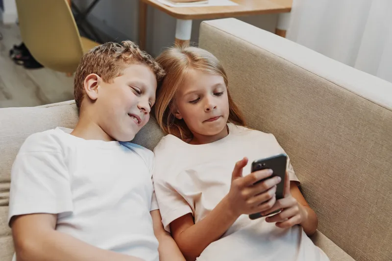 Two children sitting on a beige sofa closely watching a smartphone screen together. Casual home interior with natural light creates a relaxed and cozy atmosphere for shared entertainment.