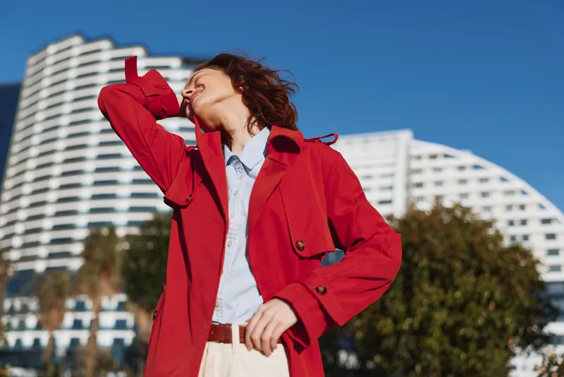 Young woman in a red coat enjoys the sunshine outdoors with eyes closed and head tilted back in a relaxed urban environment.