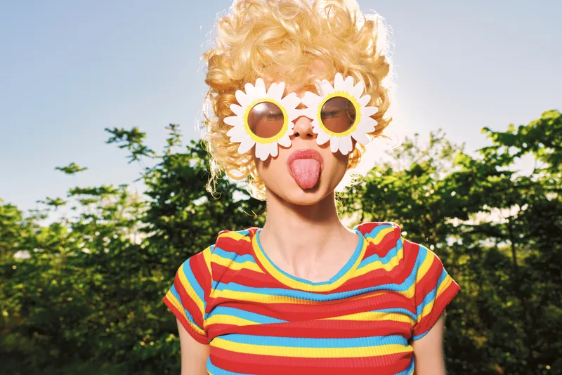 Playful young woman with blonde curls wears flower sunglasses and a striped tee in a sunlit park, sticking out her tongue for a goofy, carefree, lighthearted moment.