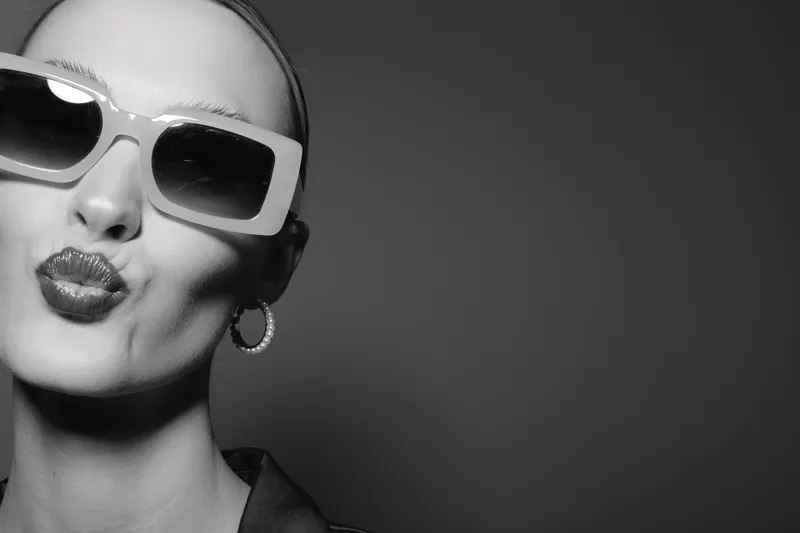 Portrait of a white woman with pale skin wearing large sunglasses and hoop earrings making a kiss face. Black and white studio photo with stylish and playful mood.