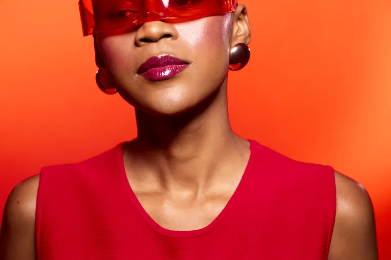 Close-up of a confident dark skinned African woman wearing red sunglasses and red dress against an orange background. Concept of fashion, style, and bold beauty with vibrant colors.