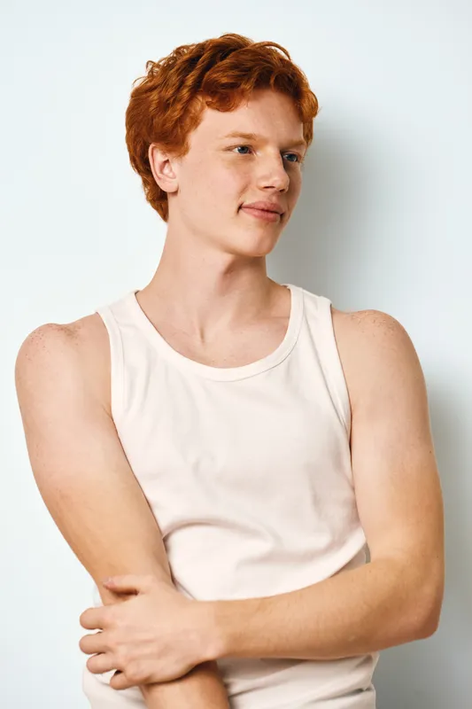 Young man with red hair and fair skin wearing white tank top standing and looking sideways against white background. Casual confident male posing with folded arms and relaxed expression.