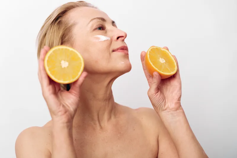 Mature woman with natural skin holds fresh orange halves near face, applying white cream on cheek. Concept for skincare, healthy lifestyle, natural beauty, and citrus wellness in studio.