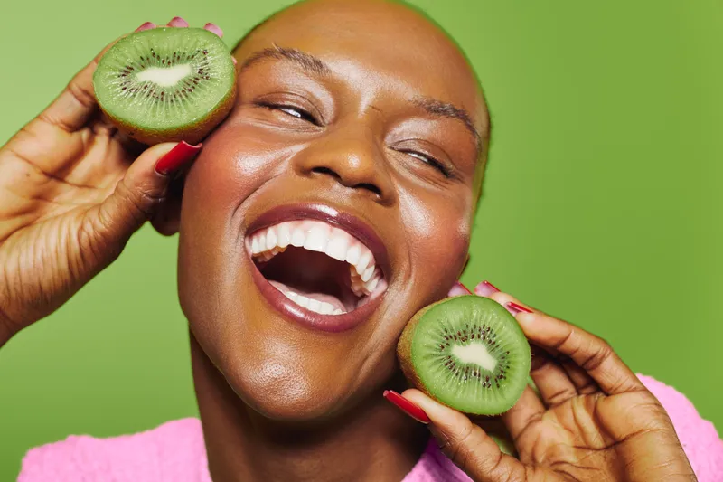 Joyful dark skinned African woman with shaved head holds two halves of fresh kiwi near face on green background. Concept of healthy food, natural beauty, skincare and positive lifestyle.