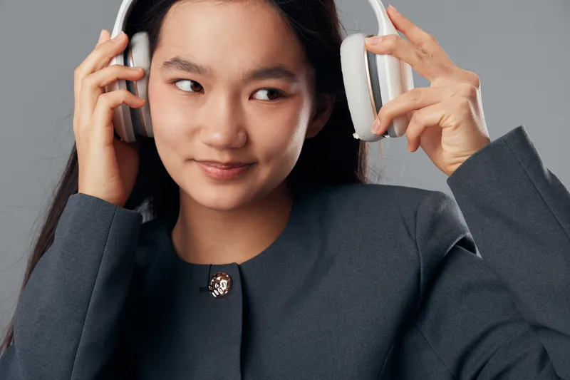 Young woman putting on headphones with smile and casual blazer on gray background. Asian female, music listening and relaxed expression for technology entertainment and lifestyle.
