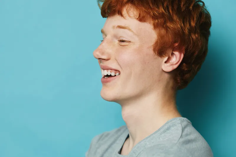 Teenager boy with red hair and freckles smiling sideways against blue background. Casual portrait with natural light and happy expression on face in studio with copy space.