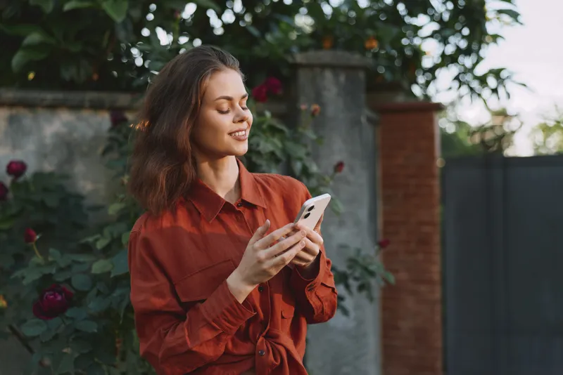 Cheerful woman outdoors, holding phone, relaxed lifestyle moment with sunshine, casual red shirt, natural garden backdrop, candid smile, gentle breeze, vibrant colors, comfortable pose