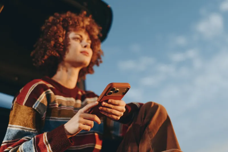 Woman lifestyle portrait outdoor, wearing hat and striped sweater, curly afro hair, holding smartphone and checking screen, relaxed pose against bright sky in daylight, closeup.