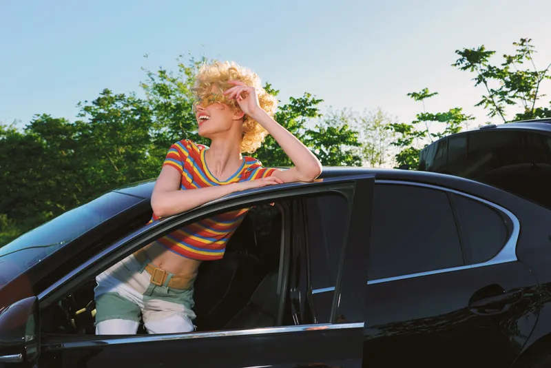 A young woman with blonde curly hair leans out of a glossy black car window, smiling in a striped crop top and denim shorts on a sunny day.