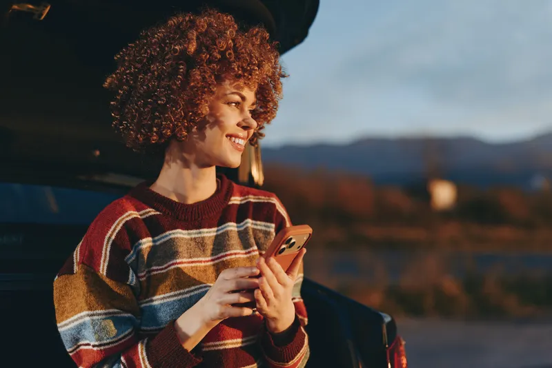 Woman lifestyle outdoors, curly hair and warm sweater, holding smartphone near face, standing by car at sunset, blurred mountains behind, candid communication, relaxed evening mood