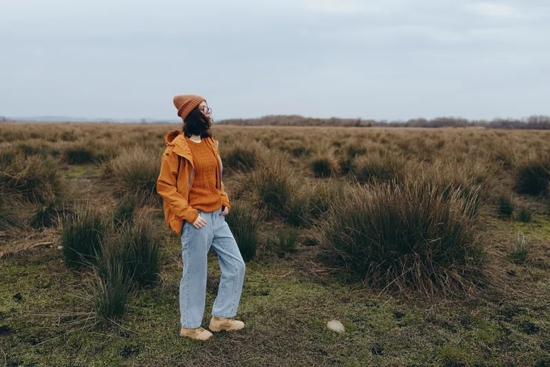 woman outdoors, field, nature, lifestyle, standing, jacket, jeans, grass, cloudy sky, orange coat, hands in pockets, looking away, calm mood, soft natural light, wide landscape