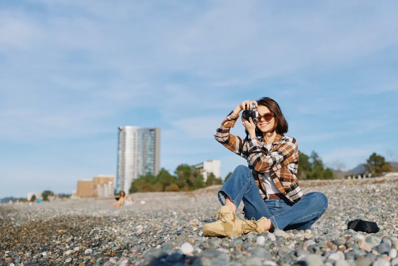 Woman outdoors on a pebble beach with a camera, dressed in plaid jacket and boots, capturing a sunny coastal landscape with an urban backdrop.