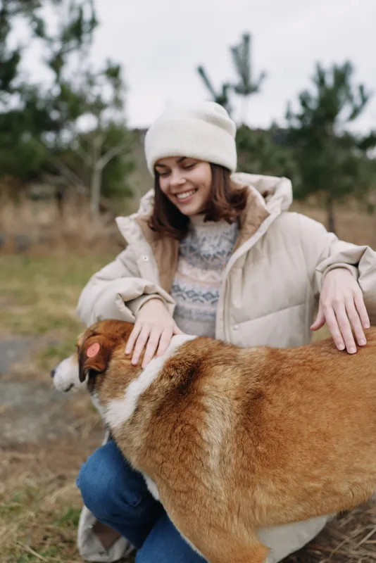 Woman kneeling beside a large dog outdoors, wearing a warm coat and beanie, sharing a gentle cuddle in a peaceful countryside setting, with grass, trees in distance and soft light