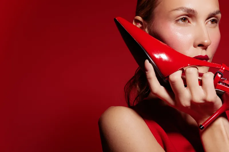 Close-up of a woman holding a red high heel shoe near her face against a red background. Concept of fashion, style, elegance, and bold color contrast in studio lighting.