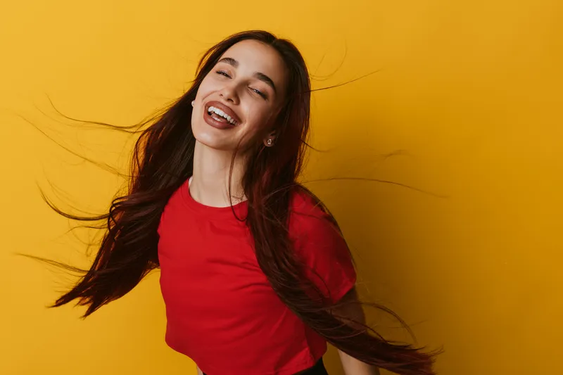 Woman smiling and laughing with long hair flowing wearing red shirt against yellow background. Happy female portrait with joyful expression and casual style in studio lighting.