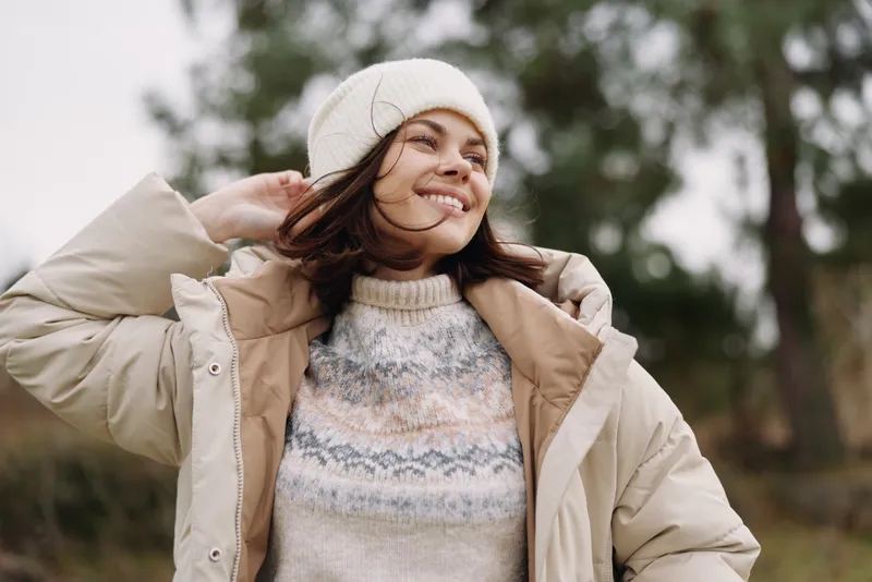 Woman outdoors wearing a beige coat and knitted sweater, smiling as wind lifts her hair in a natural park setting with soft morning light.