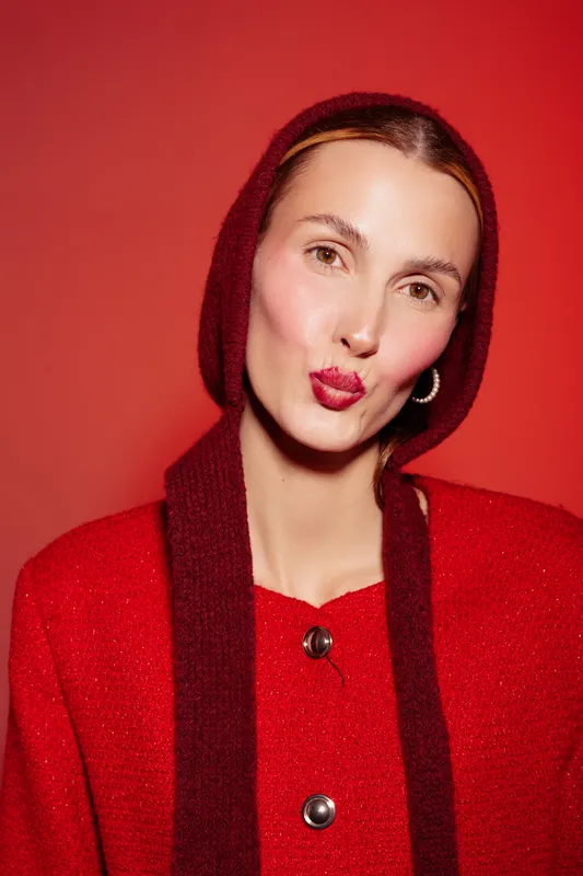 Young woman in red sweater and matching scarf makes a playful kiss face on red background. Studio portrait with bright lighting and warm tones. Concept of fun, fashion, and beauty.