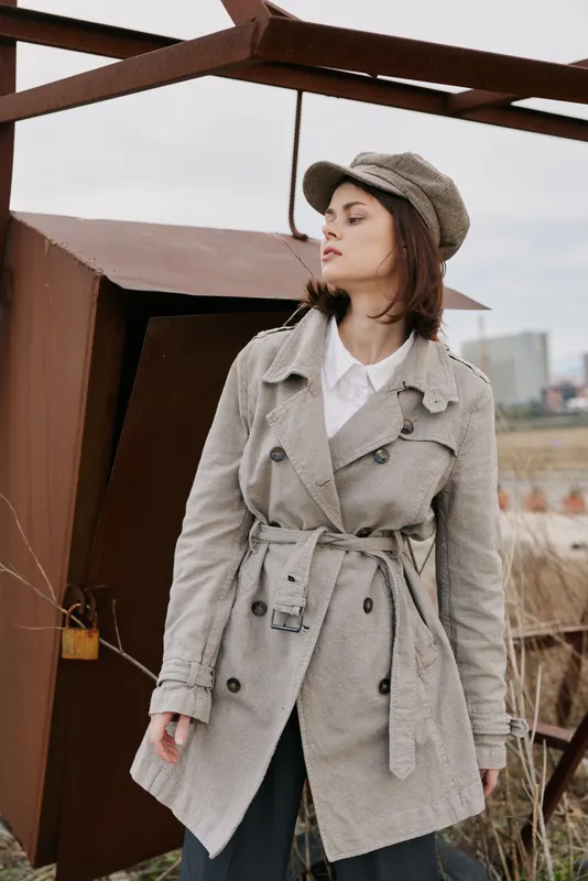 Young woman in beige trench coat and matching cap stands outdoors near rusty metal structure, looking to side with serious expression. Urban style, fashion, casual, autumn mood concept.