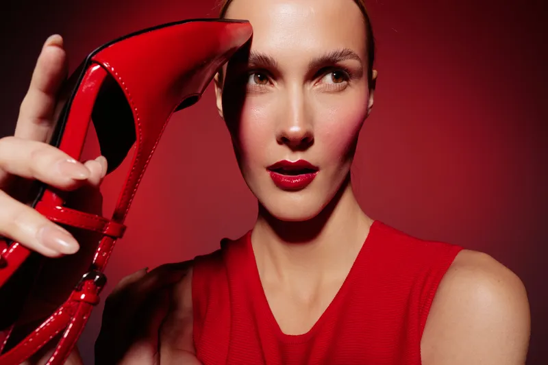 Close-up of confident woman in red dress holding red high heel shoe near face on red background. Concept of fashion, style, beauty, and elegance with bold makeup and lighting.