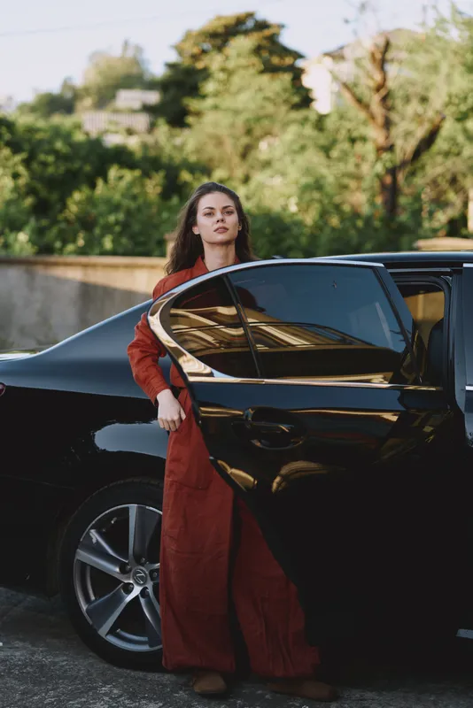 Woman in a red dress steps from a sleek black car, showcasing a refined lifestyle mood, confident pose, urban outdoor setting, soft sunlight, elevated fashion moment