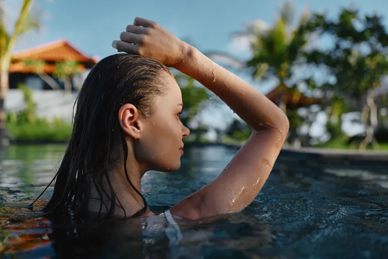 Poolside scene features a person in water, side profile with hand shielding eyes, wet hair, relaxed mood, outdoor lifestyle, leisure moment under bright sun