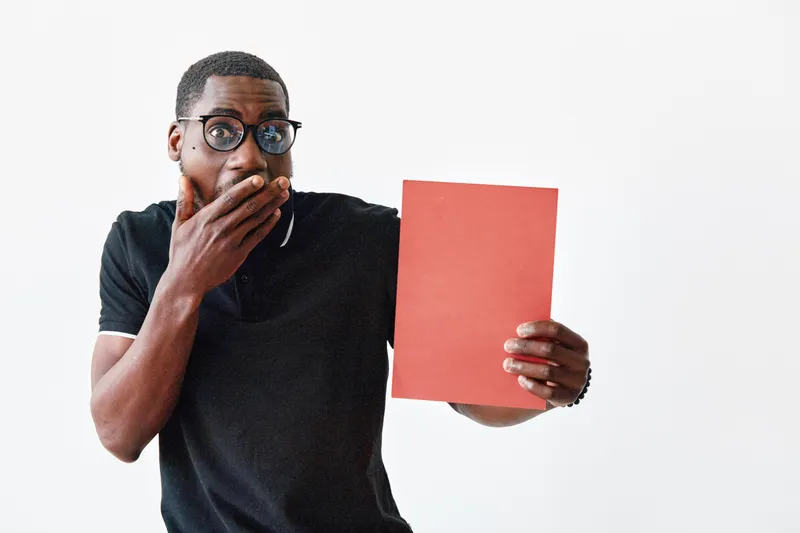 African American man with dark skin and glasses holding red paper with surprised expression on white background. Casual clothing and emotional reaction for communication or message.