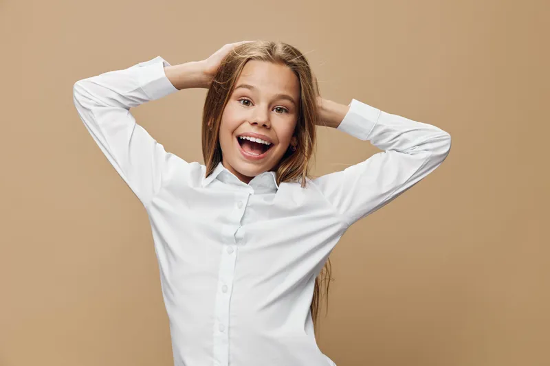 Girl with long hair smiling and posing with hands behind head on beige background. Happy child in white shirt with joyful expression and casual posture looking at camera.