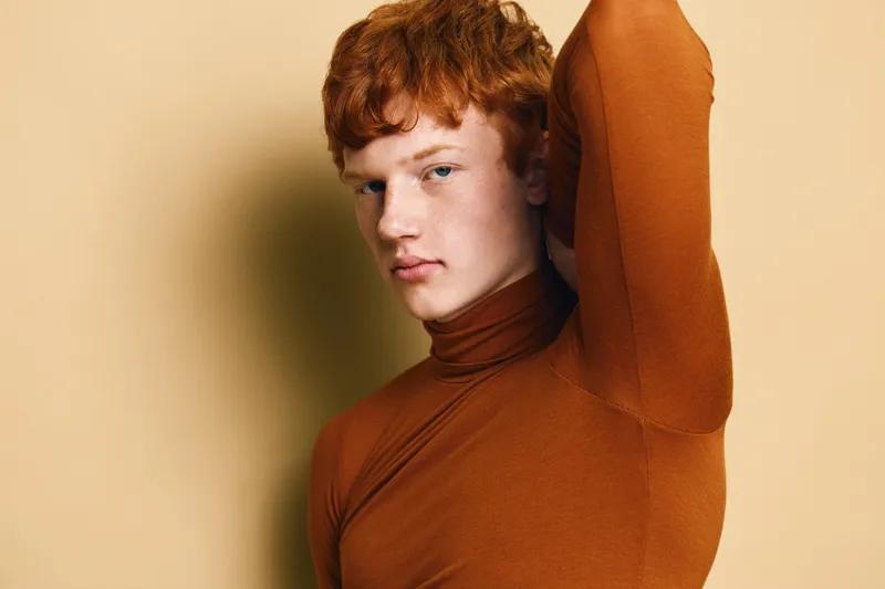 Young man with red hair and light skin wearing brown turtleneck with arm raised looking at camera against plain background in studio portrait photography.
