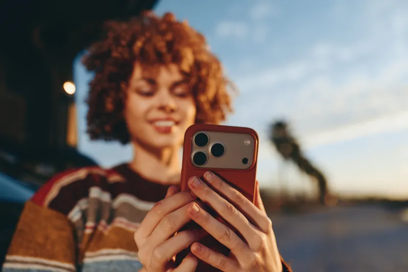 Woman lifestyle outdoors, curly hair, smiling while holding a smartphone in her hands, warm sunset light, softly blurred street background, relaxed candid moment evening breeze