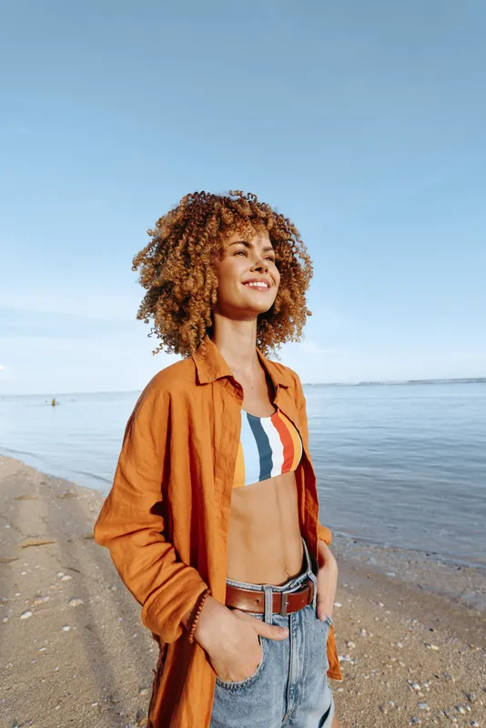 Woman at beach by sea, curly hair smiling in sunlight, outdoors with blue sky, wearing orange jacket and striped top, denim overalls, relaxed confident pose, calm horizon, view