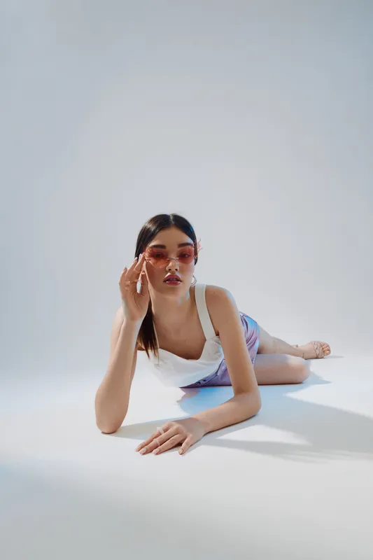Young woman in white top and shorts lying on floor wearing pink sunglasses. Minimalist studio shot with soft shadows. Concept of beauty fashion and modern style.