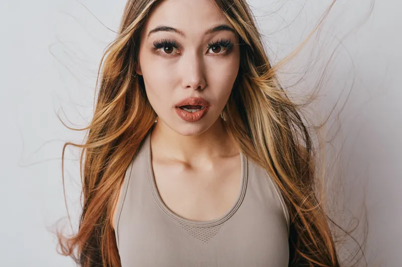 Young woman with long blond hair and natural makeup looking surprised in beige top on white background. Expression, emotion, female, beauty, casual and indoor portrait studio.