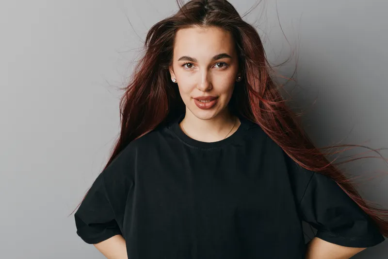 Young woman with long hair and casual black t shirt standing against gray background looking at camera with confident expression and hands on hips in studio portrait.
