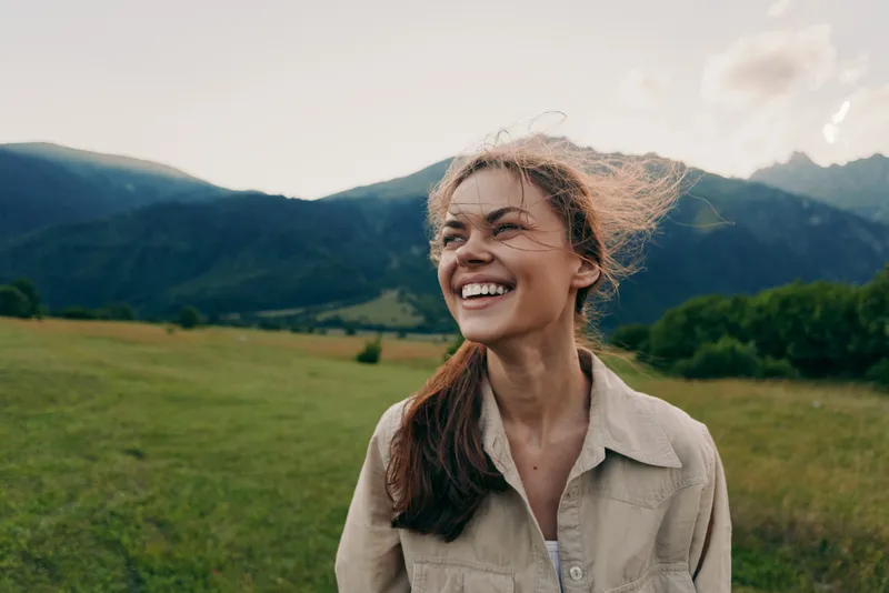 Young woman outdoors in a sunlit field with distant mountains, smiling with wind in her hair, casual beige shirt, relaxed pose and natural colors.
